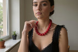 A woman wearing a chunky red beaded necklace, posing indoors near a window with plants on the sill.