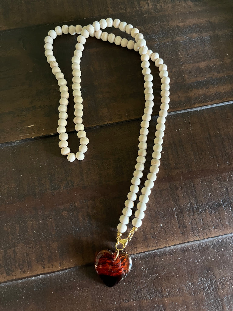A double-strand beaded necklace with small off-white beads and a heart-shaped pendant containing red and black marbled details, resting on a dark wooden surface.