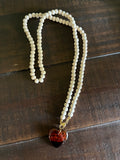 A double-strand beaded necklace with small off-white beads and a heart-shaped pendant containing red and black marbled details, resting on a dark wooden surface.
