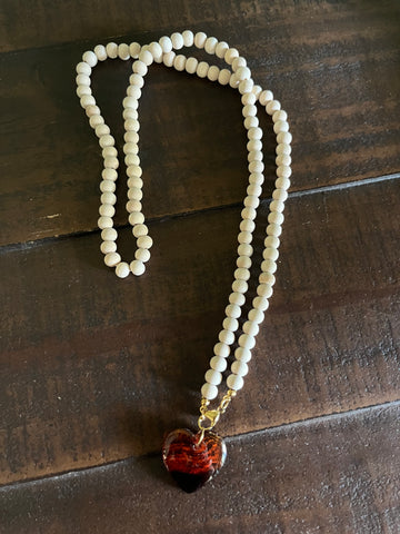 A double-strand beaded necklace with small off-white beads and a heart-shaped pendant containing red and black marbled details, resting on a dark wooden surface.