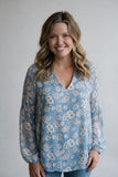 A smiling woman with blonde wavy hair wearing a light blue long-sleeve blouse with a white and pink floral pattern, paired with blue jeans, standing against a neutral light gray background.