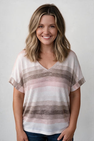 A smiling woman with shoulder-length blonde hair wearing a short-sleeve V-neck top with horizontal stripes in white, dusty pink, and taupe, paired with dark blue jeans, against a light gray studio background.