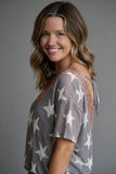 A smiling woman with shoulder-length brown hair wearing a gray short-sleeve top with white star patterns and rose gold lace shoulder detailing, photographed against a solid gray studio background.