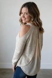 A woman wearing a light beige knit sweater with a cold-shoulder back design and a draped open back, paired with blue jeans, photographed against a neutral light gray wall.