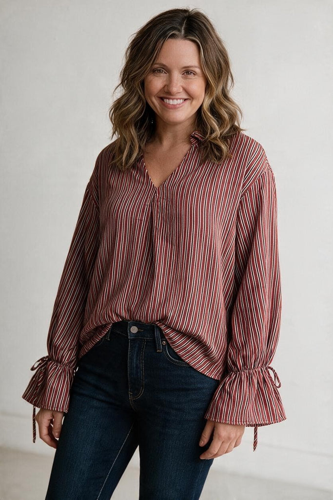 A woman wearing a long-sleeve button-up blouse with vertical red, pink, and white stripes, paired with dark blue jeans, standing against a light gray wall.
