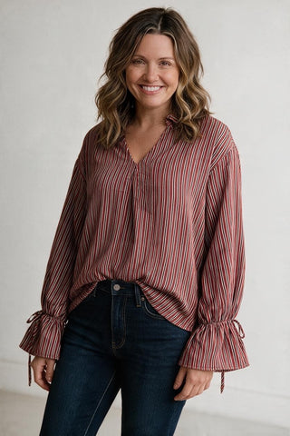 A woman wearing a long-sleeve button-up blouse with vertical red, pink, and white stripes, paired with dark blue jeans, standing against a light gray wall.