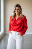 A smiling woman with blonde hair stands indoors wearing a bright red long-sleeve blouse with a draped neckline and white pants with gold buttons.