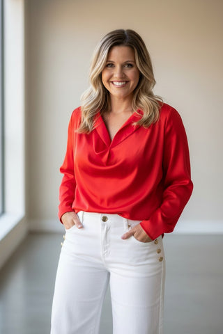 A smiling woman with blonde hair stands indoors wearing a bright red long-sleeve blouse with a draped neckline and white pants with gold buttons.