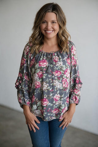 A woman smiling while wearing a gray long-sleeve blouse with a pink and white floral print, paired with blue jeans, standing against a plain light gray wall.