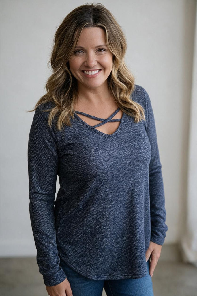 A smiling woman with blonde-highlighted hair wearing a dark heathered blue long-sleeve top with a criss-cross neckline, paired with blue jeans, standing against a neutral indoor wall.