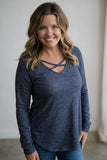 A smiling woman with blonde-highlighted hair wearing a dark heathered blue long-sleeve top with a criss-cross neckline, paired with blue jeans, standing against a neutral indoor wall.