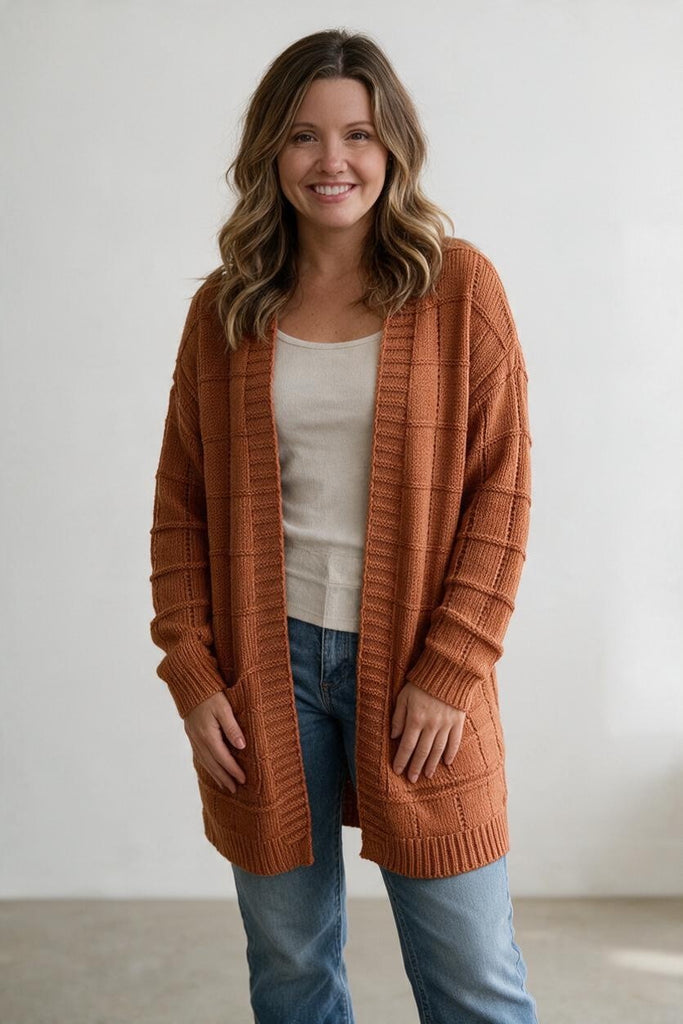 A woman wearing a rust-orange open-front cable-knit cardigan over a cream top and blue jeans, standing against a plain light gray wall.