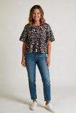 A woman modeling a short-sleeve black blouse with a beige and white floral pattern, paired with blue jeans and white sneakers, against a plain light gray studio background.