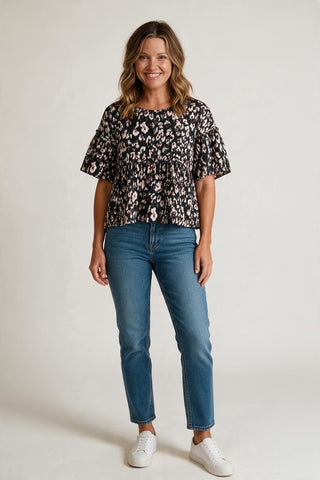 A woman modeling a short-sleeve black blouse with a beige and white floral pattern, paired with blue jeans and white sneakers, against a plain light gray studio background.