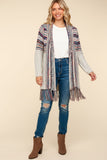 A woman modeling a long open-front cardigan with a multicolor geometric pattern, gray knit sleeves, and fringe at the hem, styled with a white t-shirt, distressed blue jeans, and brown ankle boots against a solid beige background.