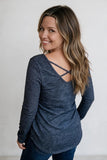 A woman with long highlighted brown hair is shown from the back wearing a dark heathered blue long-sleeve top with crisscross straps at the neckline, paired with dark blue jeans, against a plain light gray background.