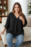 A woman wearing a loose black V-neck top and blue jeans sits on a white sofa in a bright living room.