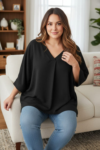 A woman wearing a loose black V-neck top and blue jeans sits on a white sofa in a bright living room.