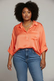 A woman with dark curly hair wearing a bright orange satin button-down shirt and blue jeans, standing against a neutral gray studio background.