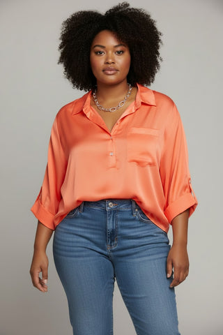 A woman with dark curly hair wearing a bright orange satin button-down shirt and blue jeans, standing against a neutral gray studio background.
