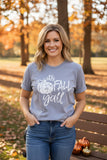 A woman wearing a heather gray graphic t-shirt with 'It's FALL y'all' and a pumpkin design, paired with blue jeans, standing in an autumn park setting.