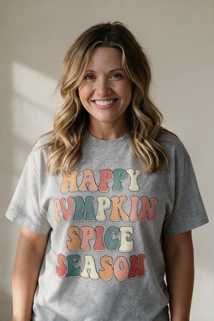 A smiling woman with wavy blonde hair wearing a heather gray graphic t-shirt that reads 'HAPPY PUMPKIN SPICE SEASON' in retro, multi-colored bubble letters, set against a warm beige background.
