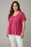 A woman modeling a short-sleeve V-neck blouse with a pink and white floral pattern, paired with white pants, against a solid gray studio background.