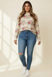 A woman modeling a beige long-sleeve floral mock-neck top with blue distressed skinny jeans and white sneakers, standing against a warm beige background with soft window shadow overlays.
