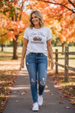 A woman walking on a path surrounded by autumn trees, wearing a white graphic t-shirt that reads 'Farm Fresh Pumpkins' with a pumpkin illustration, blue ripped jeans, and white sneakers.