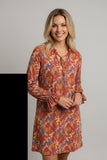 A woman modeling a long-sleeve, knee-length dress with a rust-orange base and a dense floral print in blue, yellow, and cream, featuring a tie-neck detail and ruffled cuffs, set against a plain light gray background.