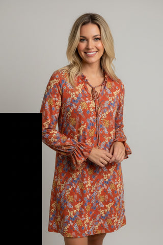 A woman modeling a long-sleeve, knee-length dress with a rust-orange base and a dense floral print in blue, yellow, and cream, featuring a tie-neck detail and ruffled cuffs, set against a plain light gray background.