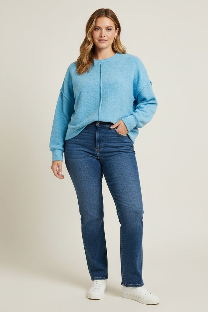 A woman modeling a light blue knit crewneck sweater and dark blue jeans with white sneakers, standing against a plain light beige studio background.