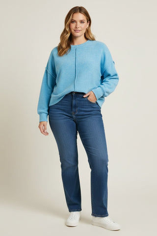 A woman modeling a light blue knit crewneck sweater and dark blue jeans with white sneakers, standing against a plain light beige studio background.