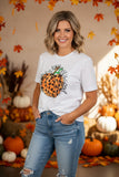 A woman wearing a white graphic t-shirt with a leopard-print pumpkin design and the text 'Happy Fall', paired with distressed blue jeans, posed in front of a warm autumn-themed backdrop with pumpkins and orange leaves.