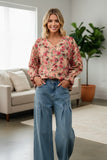 A woman stands in a bright living room wearing a long-sleeve floral blouse with a V-neck and blue wide-leg jeans.
