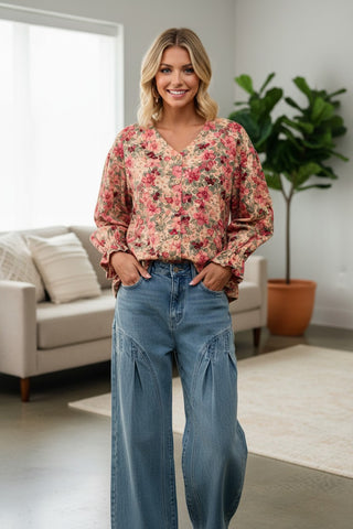 A woman stands in a bright living room wearing a long-sleeve floral blouse with a V-neck and blue wide-leg jeans.