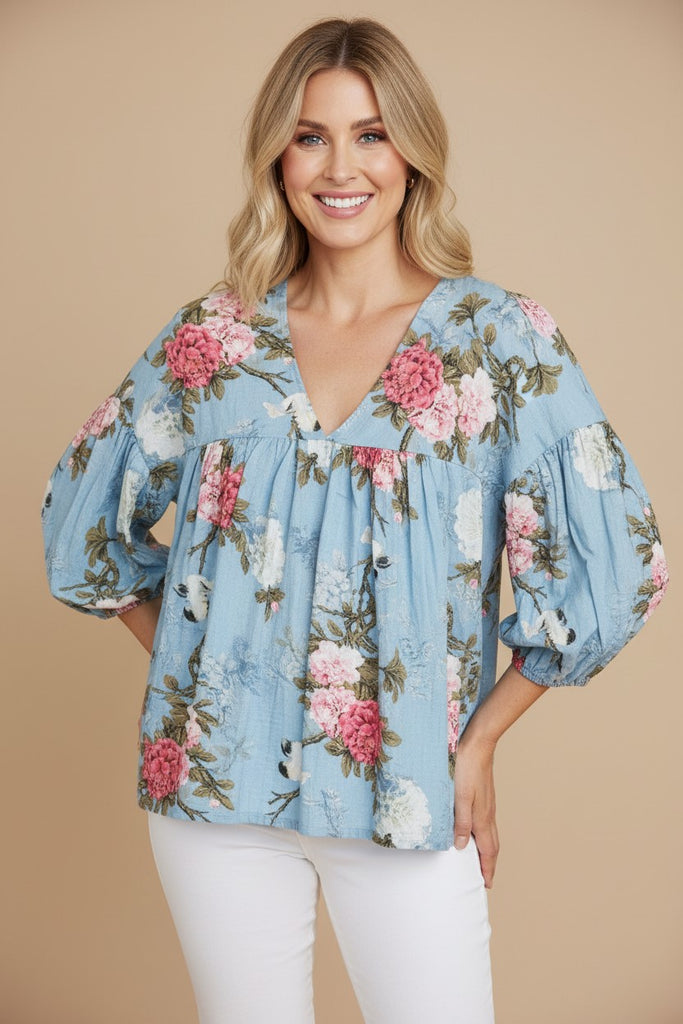A woman wearing a light blue floral blouse with puff sleeves and white pants, standing against a solid beige studio background.