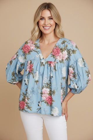 A woman wearing a light blue floral blouse with puff sleeves and white pants, standing against a solid beige studio background.