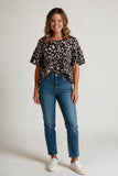 A woman modeling a black short-sleeve blouse with a white floral pattern, paired with blue jeans and white sneakers, standing against a light gray studio background.
