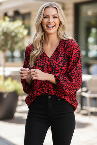 A woman with blonde hair smiling while wearing a red and black leopard print long-sleeve blouse with black pants, standing in an outdoor patio setting.