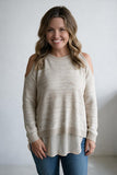 A smiling woman with wavy brown hair models a cream-colored knit sweater with cold-shoulder cutouts and a layered cream underlayer, paired with blue jeans against a neutral light gray background.