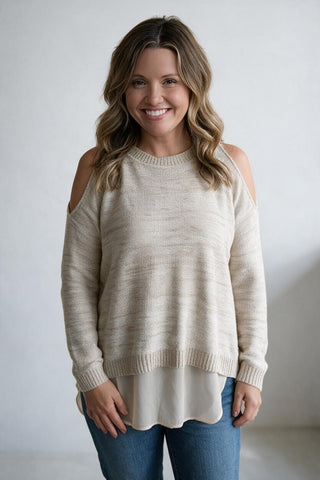 A smiling woman with wavy brown hair models a cream-colored knit sweater with cold-shoulder cutouts and a layered cream underlayer, paired with blue jeans against a neutral light gray background.