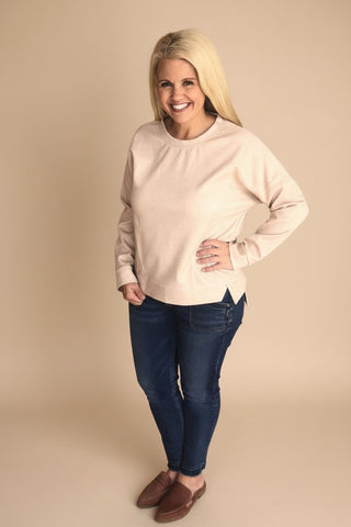 A woman modeling a light beige crewneck sweatshirt with dark blue skinny jeans and brown slip-on shoes, standing against a solid beige studio background.