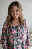 A smiling woman with wavy blonde-brown hair wearing a gray floral blouse with pink and white flowers, photographed against a soft light gray background.