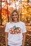 A smiling blonde woman wearing a white graphic t-shirt with an orange and white polka-dotted pumpkin design, standing in an autumn forest with colorful fall foliage.