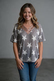 A smiling woman wearing a gray short-sleeve knit top with white star patterns and a rose gold sequined V-neck trim, paired with blue jeans, standing against a neutral gray studio background.