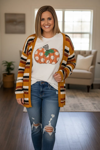 A woman stands in a living room wearing a white graphic T-shirt with a pumpkin design, layered under a mustard yellow, orange, black, and white striped cardigan, paired with distressed blue jeans.