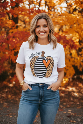 A woman smiling outdoors in front of autumn foliage, wearing a white graphic T-shirt with a striped and leopard-print pumpkin design and the words 'hello fall,' paired with blue jeans.