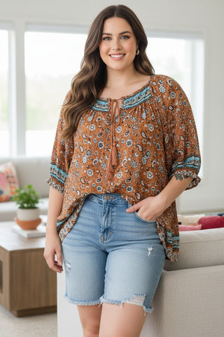 A woman modeling a brown bohemian-style blouse with a blue and white floral pattern, tie neckline, and ruffled sleeves, paired with light-wash denim shorts in a bright living room setting.