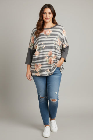 A woman modeling a gray and white striped long-sleeve top with a pink floral print and solid gray three-quarter sleeves, paired with blue jeans and white sneakers, against a solid gray studio background.